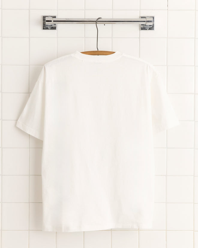 The Beaded Necklace Tee, crafted from 100% cotton, hangs on a wooden hanger against a white tiled wall, highlighting its soft and unique texture.