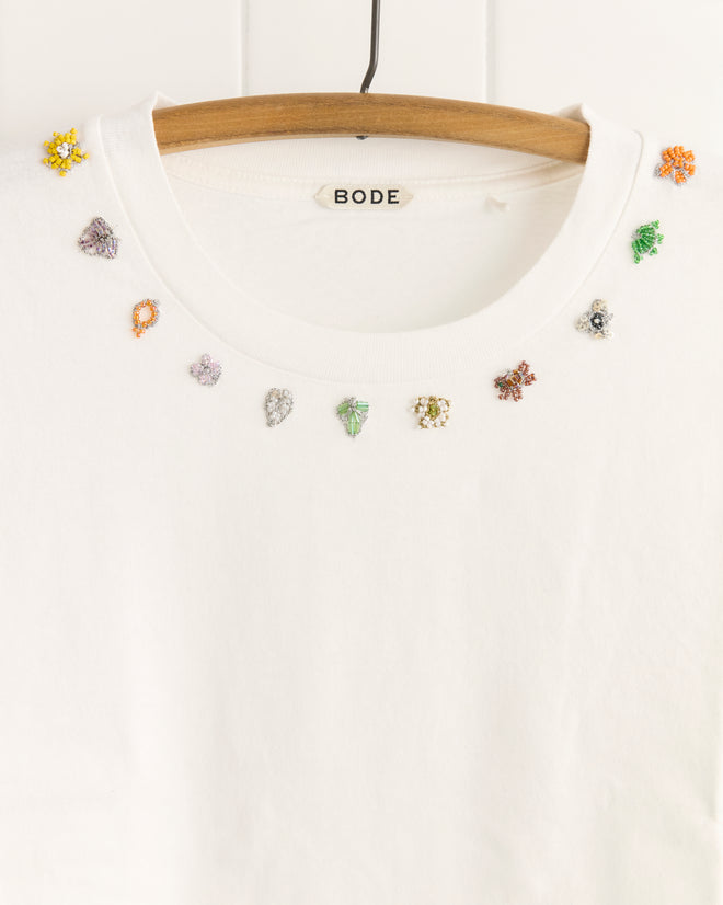 The Beaded Necklace Tee is a white Bode t-shirt on a hanger, made from 100% cotton and adorned with vibrant, beaded embroidery inspired by distinctive textile patterns around the neckline.