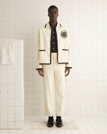 The person wearing the Beaded Player Jacket in ecru felted wool, showcasing subtle black details and a hint of a Bode logo, stands on a tiled floor, facing forward.