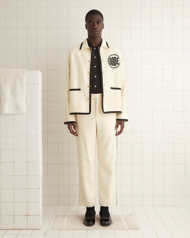 The person wearing the Beaded Player Jacket in ecru felted wool, showcasing subtle black details and a hint of a Bode logo, stands on a tiled floor, facing forward.