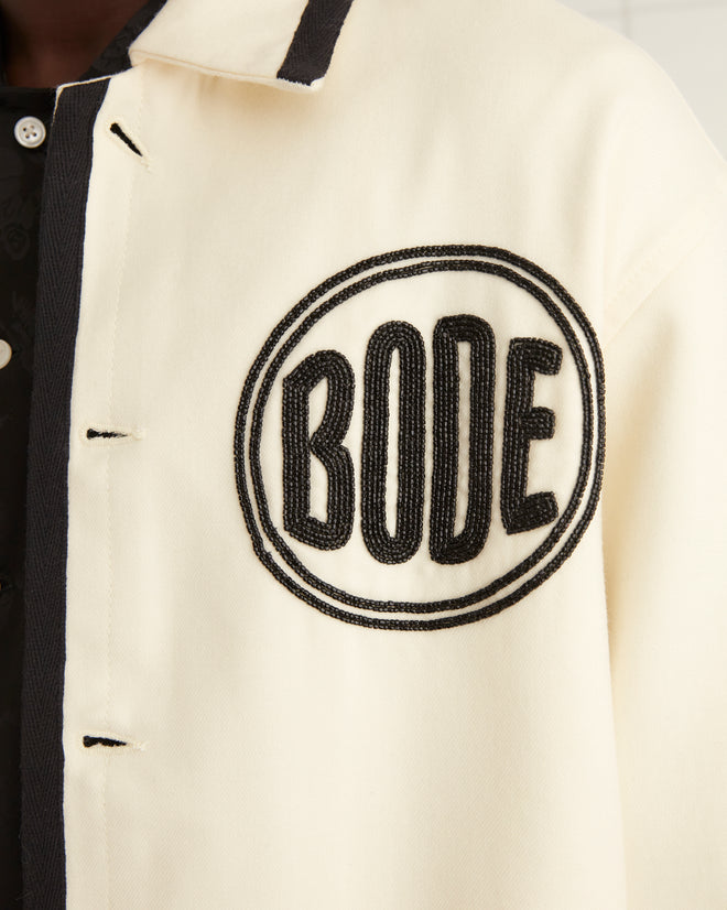 The person wearing the Beaded Player Jacket in ecru felted wool, showcasing subtle black details and a hint of a Bode logo, stands on a tiled floor, facing forward.