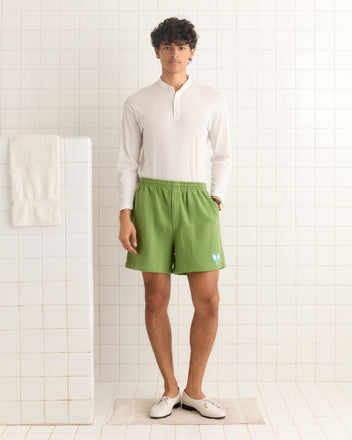 A man in a white shirt and Beaded Racquet Patch Shorts stands on a bath mat in a white-tiled bathroom.