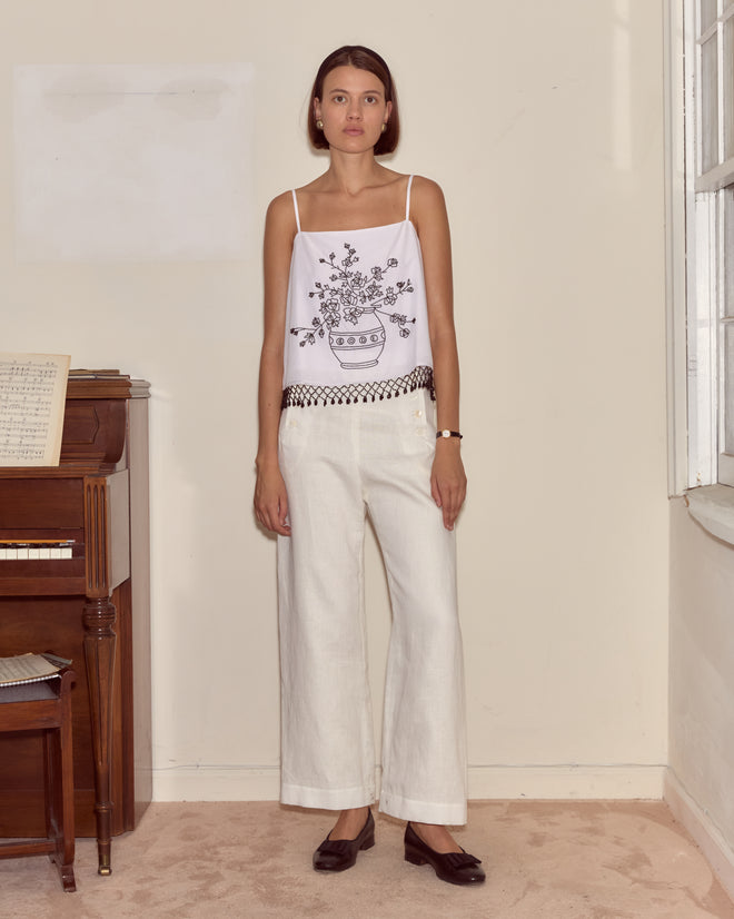 A woman in white wide-leg pants and the Beaded Rosebloom Tank stands indoors near a piano and window.
