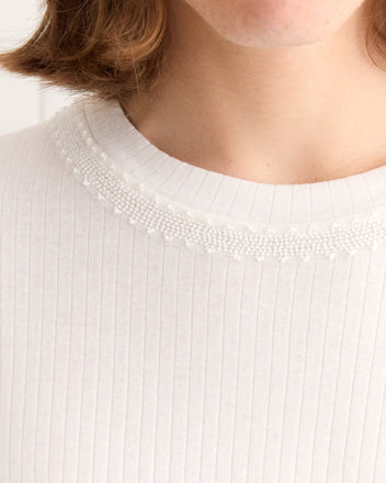 Close-up of a person wearing the Beaded Salt Marsh Tee, a tonal white ribbed cotton shirt featuring intricate beadwork around the neckline. Partial face visible.