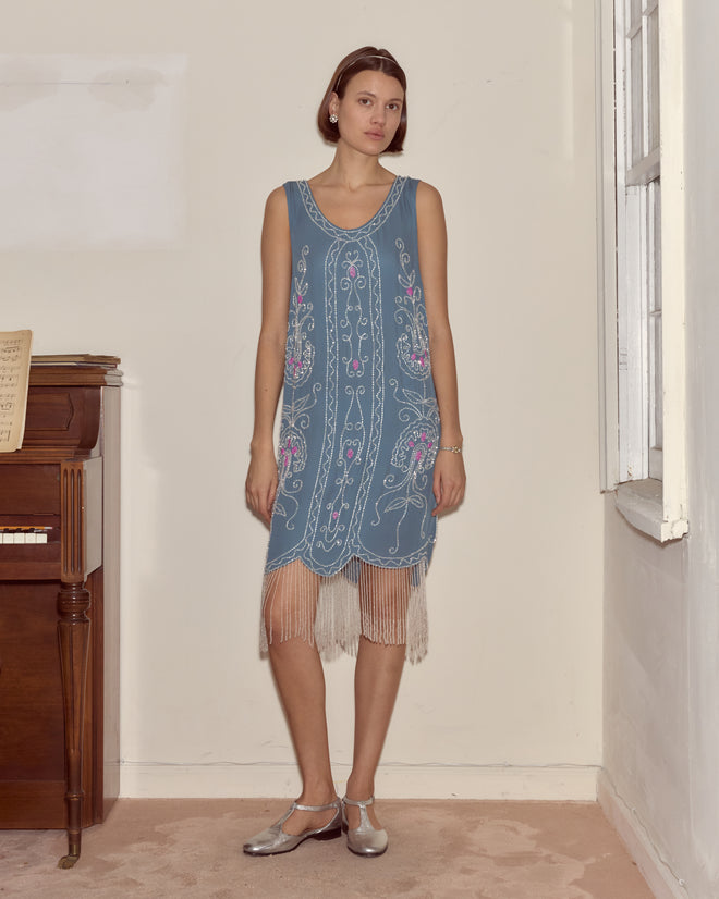 A woman in the Beaded Sea Flower Dress stands indoors by a piano and a window, looking at the camera.