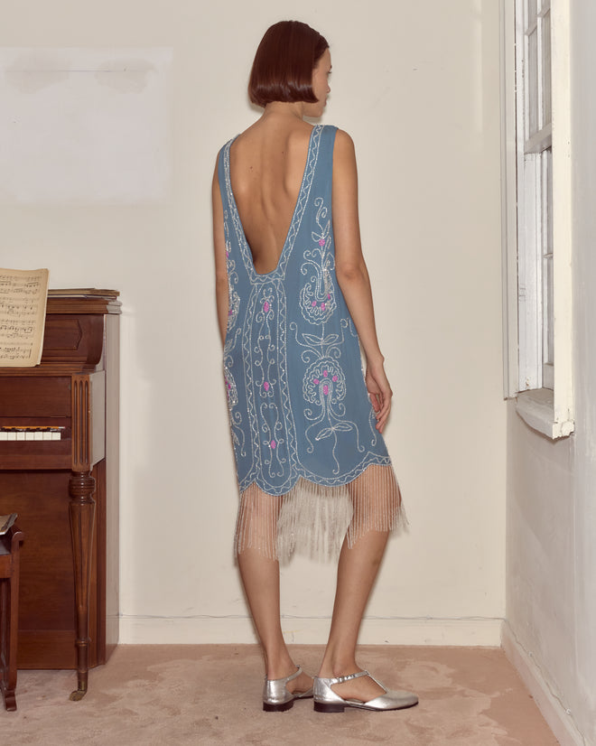 A woman in the Beaded Sea Flower Dress, a blue 1920s-inspired gown with silk fringe, stands by a piano and window, facing away.