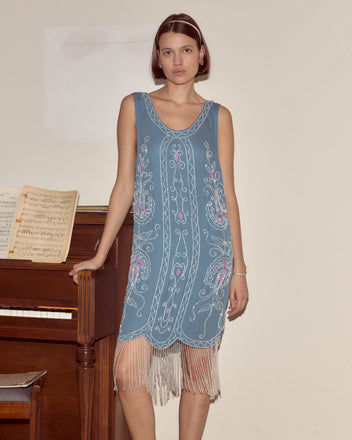 A woman in the Beaded Sea Flower Dress, a blue 1920s-style gown with a scalloped hem, stands by a piano holding sheet music against a plain wall.