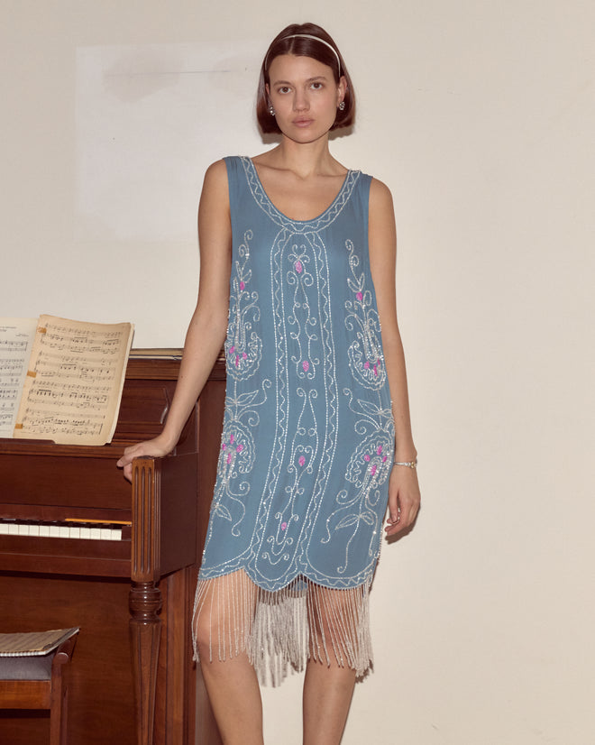A woman in the Beaded Sea Flower Dress, a blue 1920s-style gown with a scalloped hem, stands by a piano holding sheet music against a plain wall.