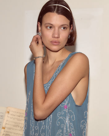 A woman in the Beaded Sea Flower Dress, featuring blue silk and a scalloped hem, adjusts her earring with sheet music visible in the background.