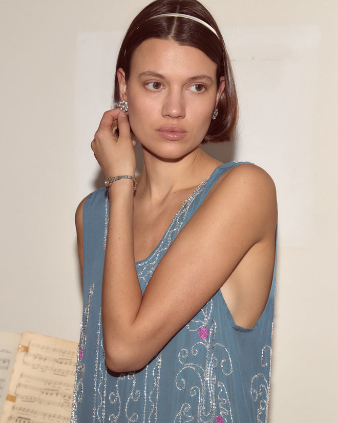 A woman in the Beaded Sea Flower Dress, featuring blue silk and a scalloped hem, adjusts her earring with sheet music visible in the background.