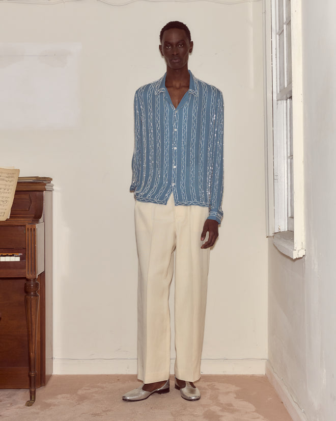 Wearing the Beaded Sea Stripe Shirt and cream trousers, a person stands indoors near a piano and a window.