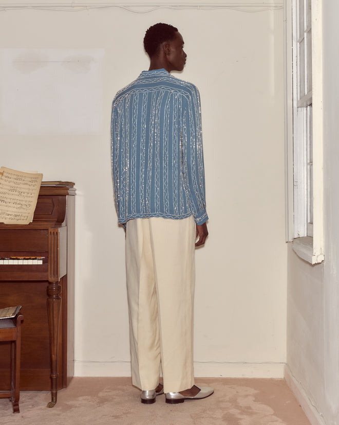 A person wearing a Beaded Sea Stripe Shirt and cream pants stands facing a wall near a piano and sheet music.