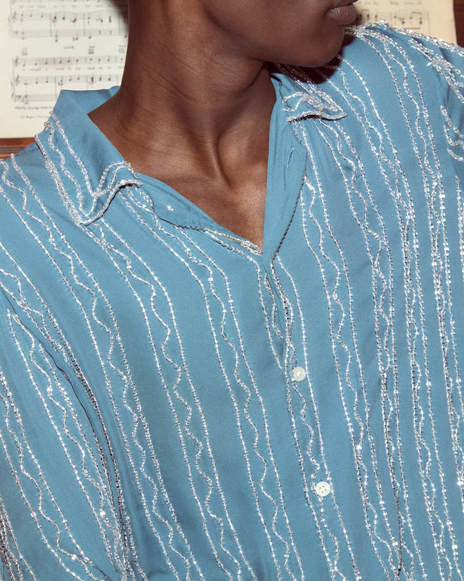 A person wearing the Beaded Sea Stripe Shirt, ocean blue with silver stripes, stands in front of a sheet of music.