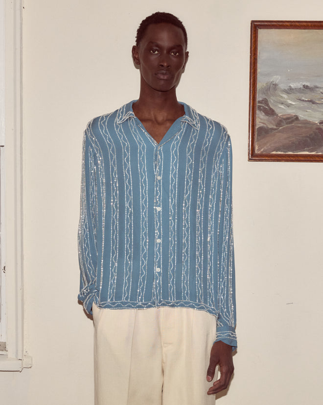 A person wearing a Beaded Sea Stripe Shirt with cream pants stands indoors beside a framed ocean painting.