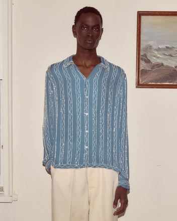 A person wearing a Beaded Sea Stripe Shirt with cream pants stands indoors beside a framed ocean painting.