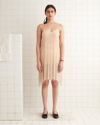 In a room with tiled flooring, a woman gracefully wears the Beaded Shakey Dress in beige, complemented by black shoes.