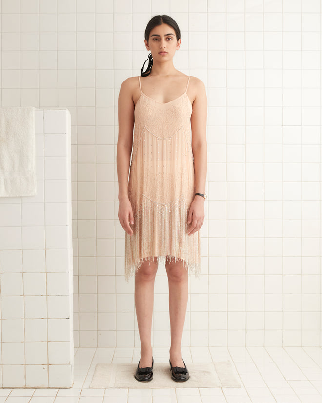 A person wearing the Beaded Shakey Dress, a beige fringe outfit inspired by 1920s beaded designs, paired with black shoes stands in a tiled room.