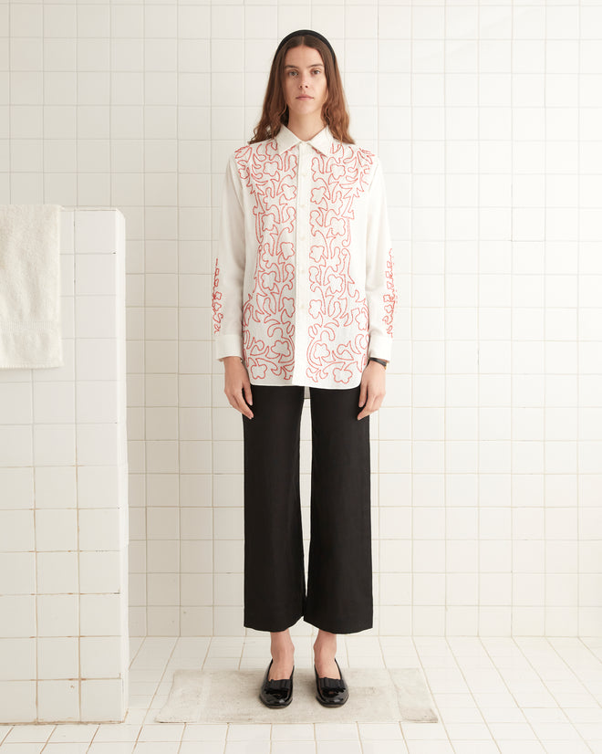 A woman stands in a tiled room wearing a Beaded Crossvine Shirt, which is a vintage white long-sleeved blouse with red patterns, black pants, and black shoes. A white towel hangs to her left.