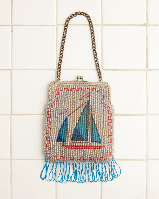 The Beaded Voyage Bag, adorned with a vintage sailboat design, features a metal chain strap and blue tassels, hanging elegantly against a tiled wall.