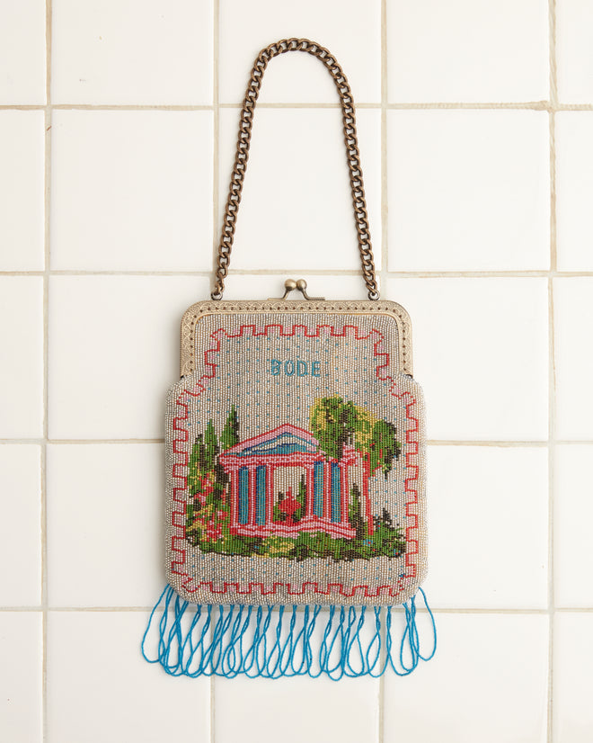 The Beaded Voyage Bag, adorned with a vintage sailboat design, features a metal chain strap and blue tassels, hanging elegantly against a tiled wall.