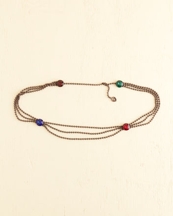 The Beadshot Gem Belt, a multi-strand bronze chain adorned with oversized gemstones in red, green, and blue, is laid out on a light beige surface.