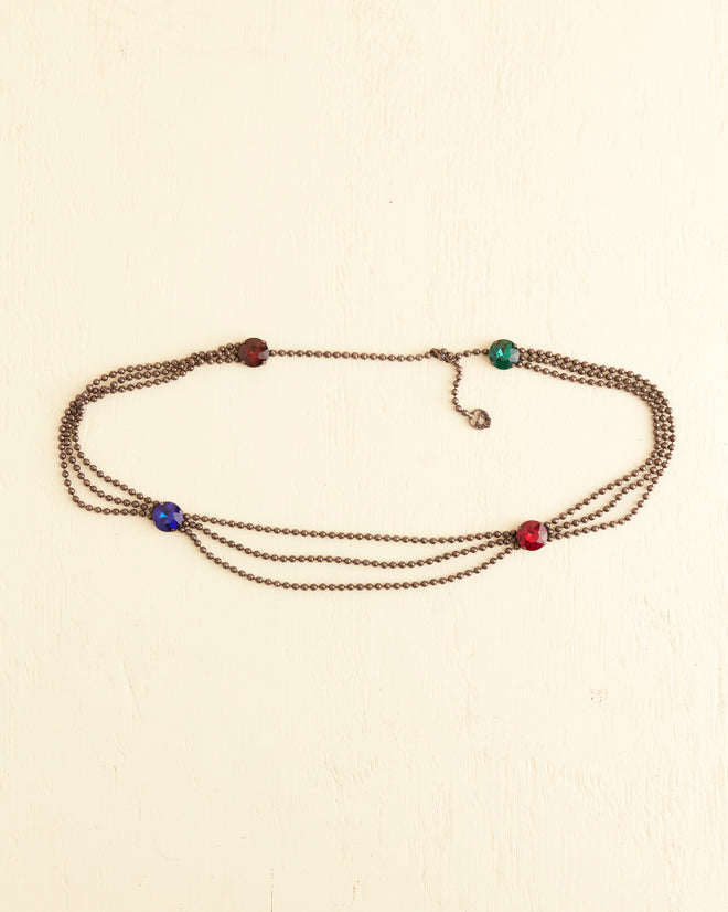 The Beadshot Gem Belt, a multi-strand bronze chain adorned with oversized gemstones in red, green, and blue, is laid out on a light beige surface.