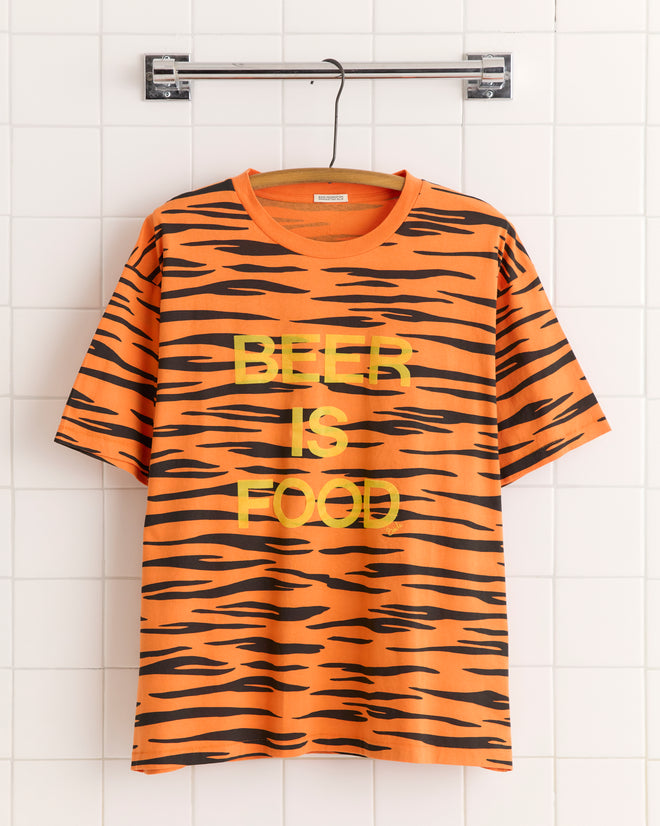 The Tiger Stripe Tee, made from 100% cotton and featuring orange tiger stripes with "BEER IS FOOD" text, hangs on a hanger against a white tiled wall.