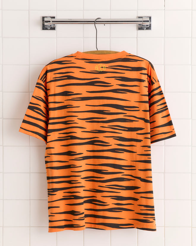 The Tiger Stripe Tee, made from 100% cotton with bold black stripes on orange, hangs on a wooden hanger against a white tiled wall.