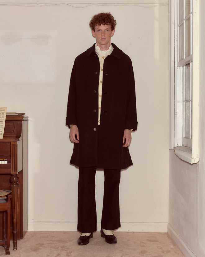 A man with curly hair stands indoors by a piano and window, wearing the Bell Coat with matching black pants.