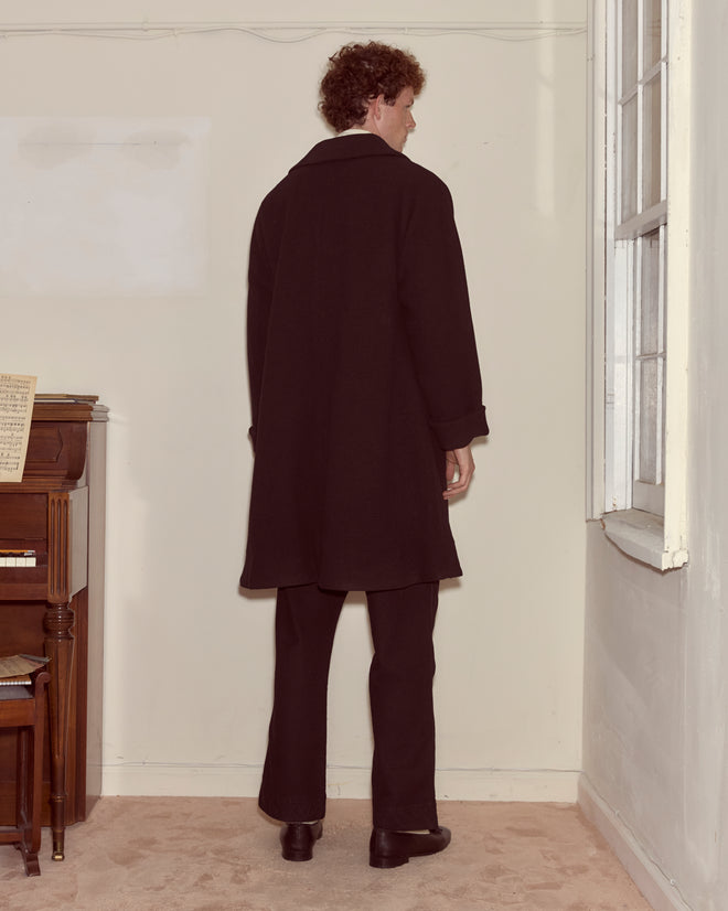 A person wearing the Bell Coat, a black wool overcoat, stands facing away in a corner near a piano and a window.