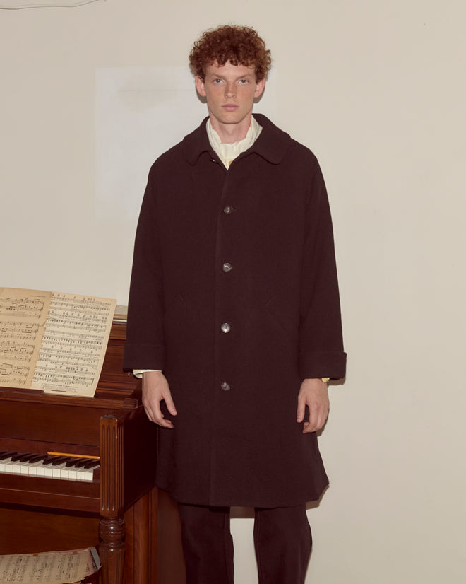 Wearing the Bell Coat, a person stands by a piano with sheet music, their silhouette cast against a plain light-colored wall.