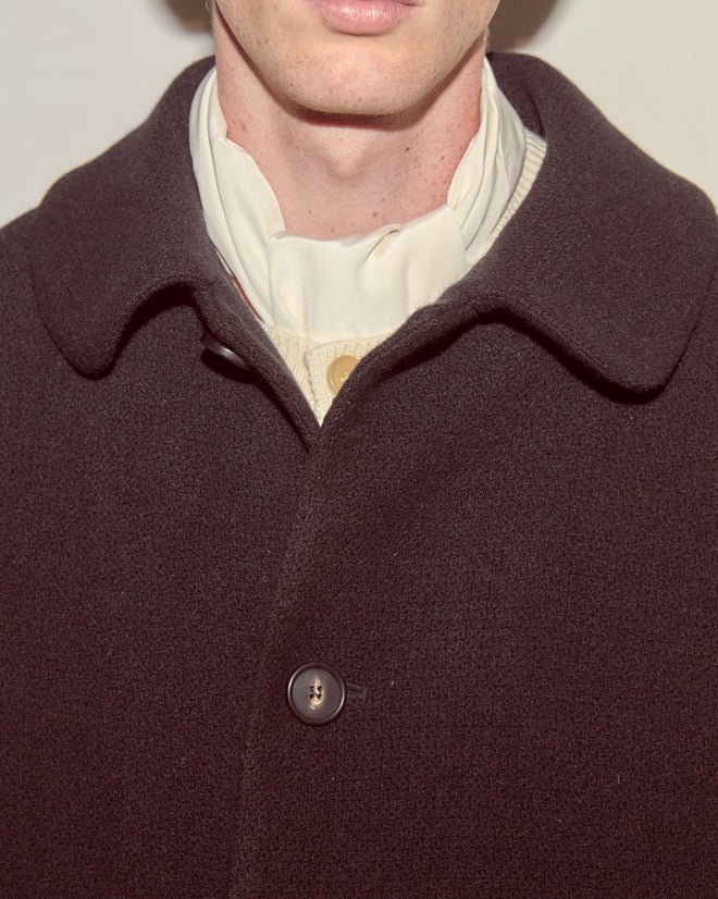 A person wearing the Bell Coat—a black wool overcoat with a swing silhouette—layered over a light shirt and finished with a cream scarf tied at the neck.