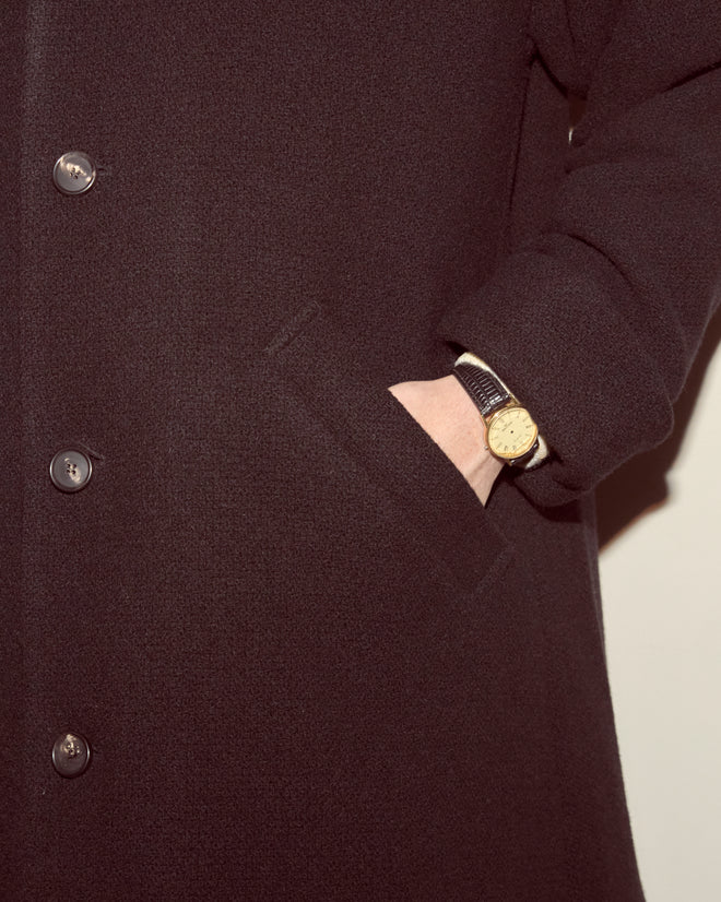 A person wears the Bell Coat, a black wool overcoat with a swing silhouette, one hand in their pocket and a gold wristwatch on their wrist—a refined statement in men's outerwear.