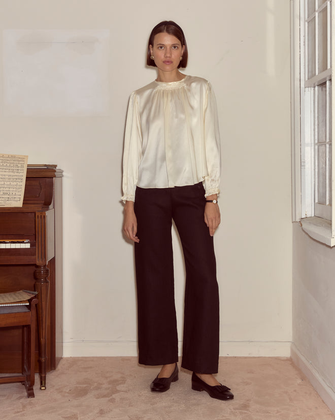A woman in the Belle Blouse - Cream, featuring traditional shirring, stands indoors beside a piano and a window, wearing black pants.