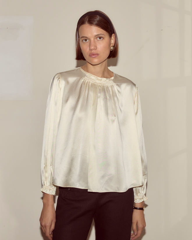 A woman wearing the Belle Blouse in cream, featuring a shiny silk finish and traditional shirring, pairs it with black pants and stands against a plain beige wall, looking at the camera.