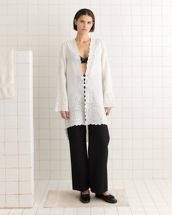 A woman in the Belvoir Top, resembling an elegant 1890s broderie anglaise bed jacket, stands gracefully in a white-tiled room, paired with sleek black pants.