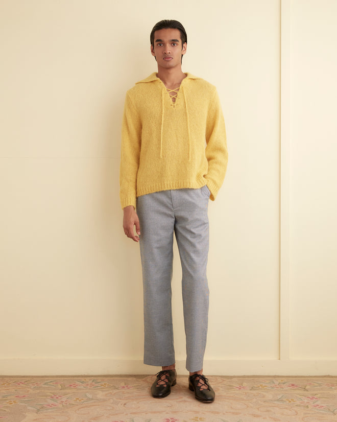 A person stands against a plain wall wearing a yellow laced-up sweater, light blue Beluga Check Trousers - 31 made from Savile Row fabrics, and black shoes. Their hands are by their sides.