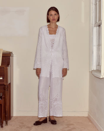 A woman wearing the Belvoir Top in white embroidered broderie anglaise stands indoors by a window and upright piano, against neutral walls.