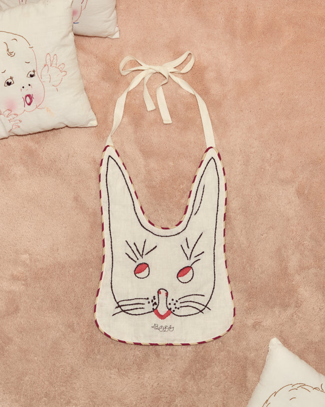 A Children's Bunny Bib rests on a soft pink surface, surrounded by hand-embroidered linen pillows that capture the whimsical charm of Alice Through the Looking Glass.
