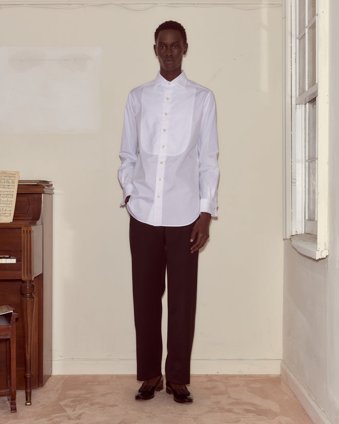 Indoors by a piano and window, a person wears the Bib Tuxedo Shirt featuring a piqué cotton bib and spread collar, paired with black pants.