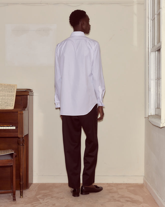 A person wearing a Bib Tuxedo Shirt with a spread collar and piqué cotton bib, paired with black pants, stands indoors facing away from the camera near a piano.