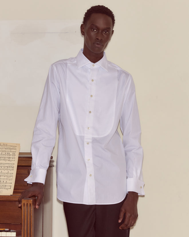 A person wearing a Bib Tuxedo Shirt with a piqué cotton bib and spread collar stands by a piano with sheet music on it.