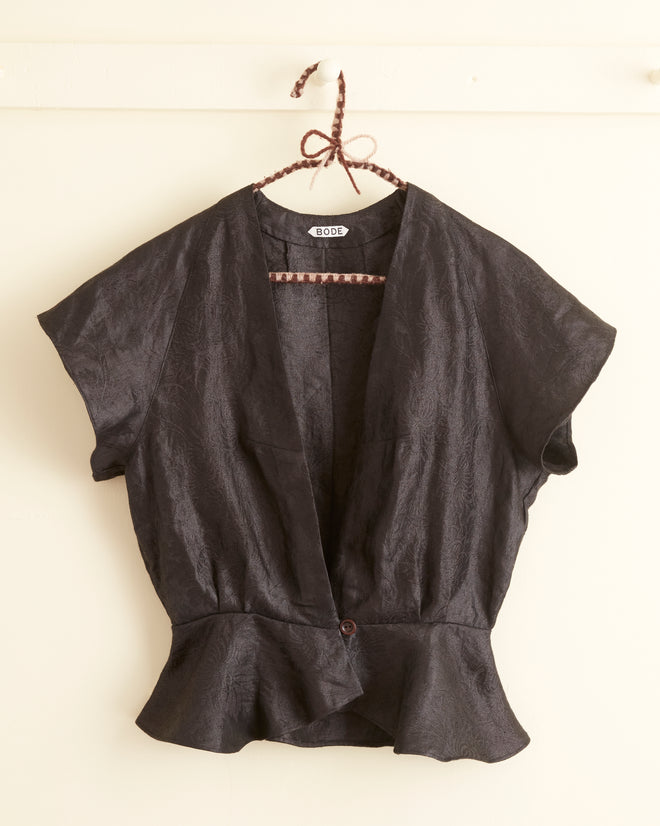 The Black Lotus Top - 2, a short-sleeved, open-front black blouse with a single button at the bottom, hangs on a wall, reminiscent of early 20th-century elegance.