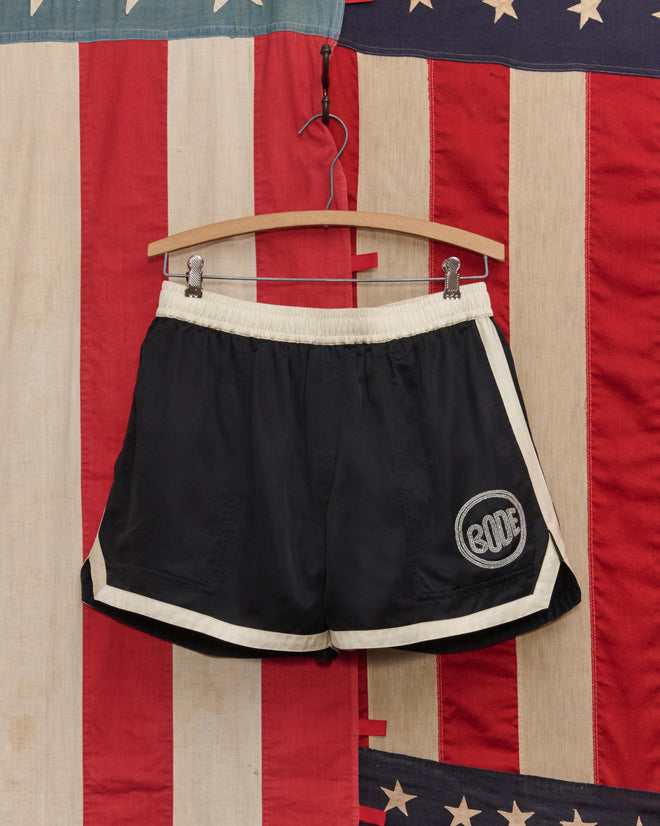 The Satin Gym Shorts - Black/Cream hang on a wooden hanger in front of an American flag backdrop, inspired by 1960s reproduction styles.