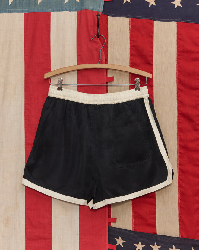 The Satin Gym Shorts - Black/Cream hang on a wooden hanger with an American flag backdrop, evoking a 1960s-inspired look.