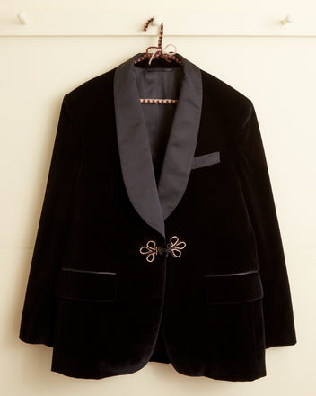 The Black Tulip Tuxedo Jacket, featuring a shawl collar, two front pockets, and a beaded frog closure, hangs on a white wall hook.