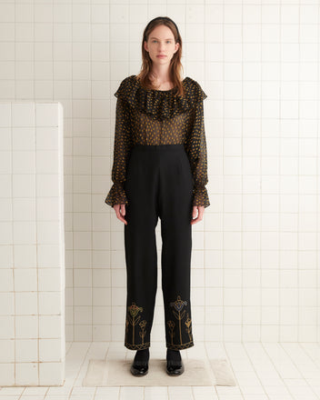 A person stands against a plain background, wearing a beige long-sleeved top and the Blooming Stems Trousers, which feature intricate black embroidery reminiscent of 1920s fashion. They have one hand in their pocket and are looking straight ahead.