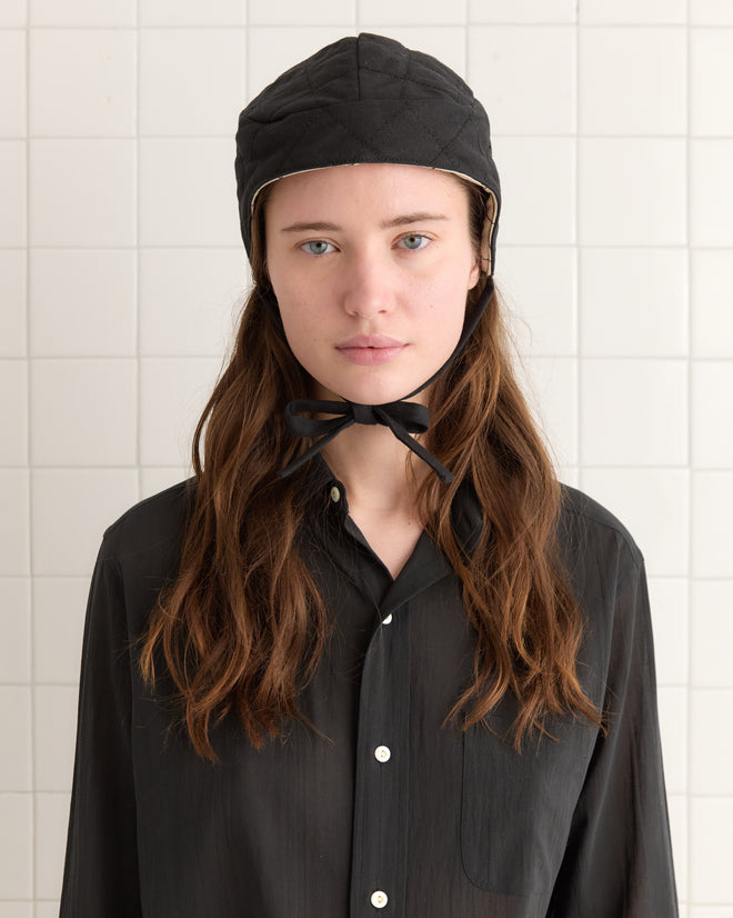 A person with long hair in a black shirt and the Bloomstein Quilted Cap stands against a tiled white wall.