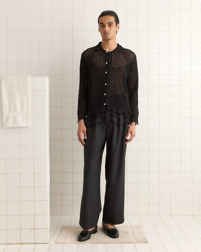 A person in a Blossom Crochet Shirt - Black and trousers stands in a tiled room, with an inviting 100% cotton towel draped nearby.