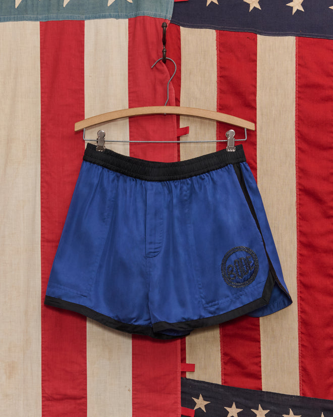 Satin Gym Shorts - Blue/Black hang on a wooden hanger in front of an American flag backdrop.